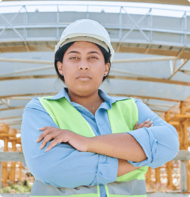 A woman wearing safety gear (hard hat, safety vest) crosses her arms in front of a building.